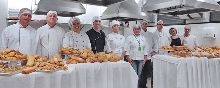 Alunos e docente do curso posam atrás de duas mesas cobertas de pães variados produzidos na unidade. Alunos e docente do curso posam atrás de duas mesas cobertas de pães variados produzidos na unidade.