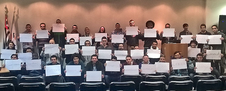 Foto de alunos enfileirados em auditório, segurando cartazes brancos com palavras como Experiência, Evolução, Foco, Dedicação Foto de alunos enfileirados em auditório, segurando cartazes brancos com palavras como Experiência, Evolução, Foco, Dedicação