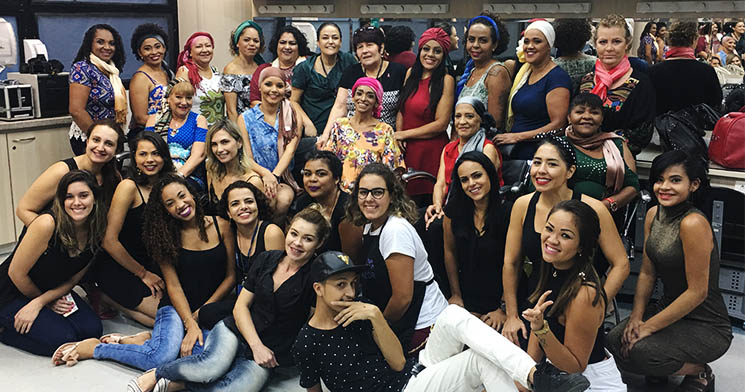 Grupo de alunas do curso Maquiador do Senac Largo Treze sentadas no chão em fileira com pacientes da ONG Amor Rosa, enfeitadas com lenços e bandanas e com maquiagens profissionais Grupo de alunas do curso Maquiador do Senac Largo Treze sentadas no chão em fileira com pacientes da ONG Amor Rosa, enfeitadas com lenços e bandanas e com maquiagens profissionais