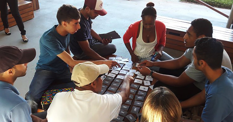 Alunos do programa Aprendizagem sentados ao redor de mesa com jogo de cartas que aborda questões de saúde e segurança do trabalho Alunos do programa Aprendizagem sentados ao redor de mesa com jogo de cartas que aborda questões de saúde e segurança do trabalho