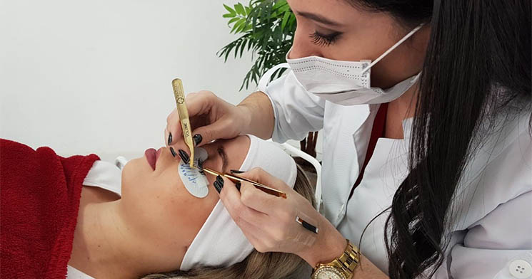 Mulher, com máscara de proteção de pano, colocando a extensão de cílios em uma cliente, deitada na maca Mulher, com máscara de proteção de pano, colocando a extensão de cílios em uma cliente, deitada na maca