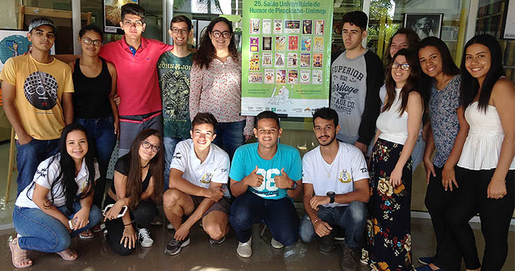 Jovens aprendizes enfileirados e sorridentes ao redor do cartaz que anunciava exposição Jovens aprendizes enfileirados e sorridentes ao redor do cartaz que anunciava exposição