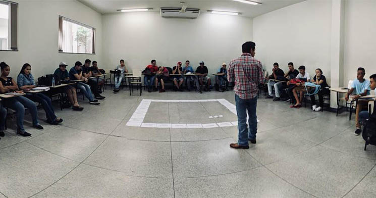 Imagem geral da sala com jogo de tabuleiro montado no chão, com homem em pé falando e jovens sentados ao redor da sala Imagem geral da sala com jogo de tabuleiro montado no chão, com homem em pé falando e jovens sentados ao redor da sala