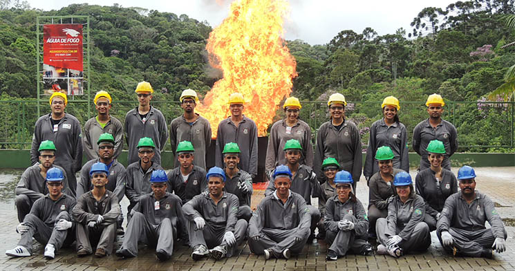 A frente de uma chama de fogo, dois homens e uma mulher posam lado a lado com macacão cinza, óculos transparente e capacete amarelo A frente de uma chama de fogo, dois homens e uma mulher posam lado a lado com macacão cinza, óculos transparente e capacete amarelo
