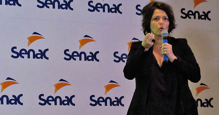 Patrícia Weiss, mulher branca de cabelos castanhos curtos, roupa social preta, em meio à fala, segurando microfone com uma mão e gesticulando com a outra mão em frente à painel branco com a logo do Senac Patrícia Weiss, mulher branca de cabelos castanhos curtos, roupa social preta, em meio à fala, segurando microfone com uma mão e gesticulando com a outra mão em frente à painel branco com a logo do Senac