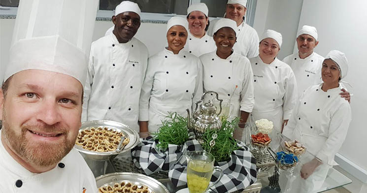 Turma com uniformes brancos reunida atrás de mesa com comidas e bebidas Turma com uniformes brancos reunida atrás de mesa com comidas e bebidas