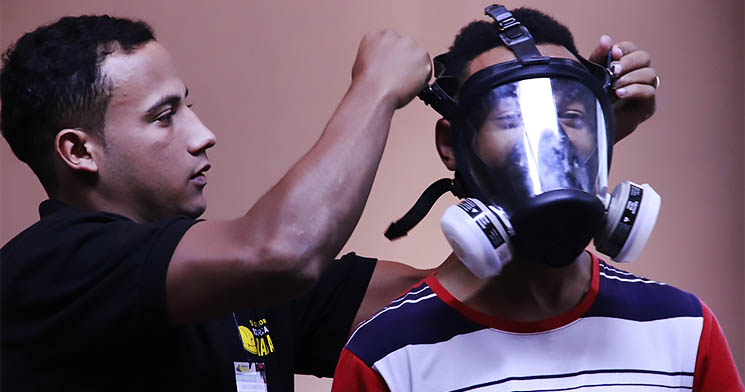Homem coloca máscara no rosto de outro homem como prevenção a acidente de trabalho Homem coloca máscara no rosto de outro homem como prevenção a acidente de trabalho