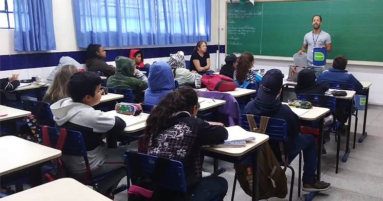 Crianças sentadas em carteiras de sala de aula de escola municipal observam apresentação de aluno do Senac São Carlos sobre coleta seletiva Crianças sentadas em carteiras de sala de aula de escola municipal observam apresentação de aluno do Senac São Carlos sobre coleta seletiva