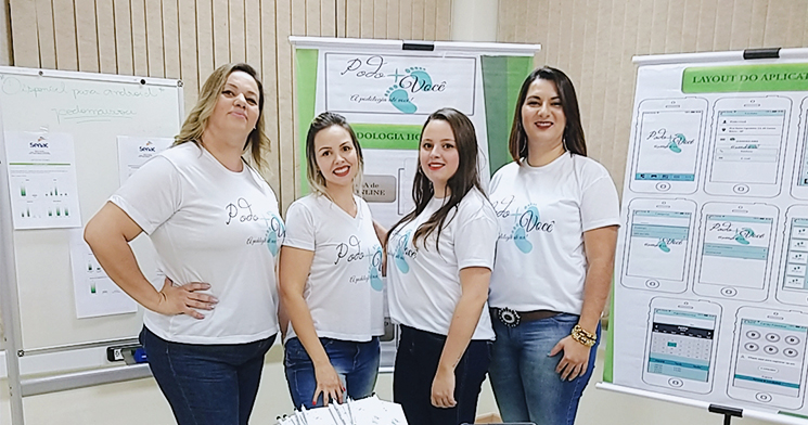 Quatro alunas do Técnico em Podologia vestindo camisetas com a logo do aplicativo em frente a banners com informações sobre o aplicativo Quatro alunas do Técnico em Podologia vestindo camisetas com a logo do aplicativo em frente a banners com informações sobre o aplicativo