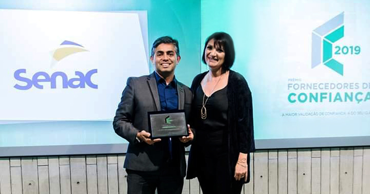 Foto de Maurício Pedro, do Atendimento Corporativo do Senac, recebendo prêmio de uma mulher, com logomarcas do prêmio e do Senac na tela ao fundo Foto de Maurício Pedro, do Atendimento Corporativo do Senac, recebendo prêmio de uma mulher, com logomarcas do prêmio e do Senac na tela ao fundo
