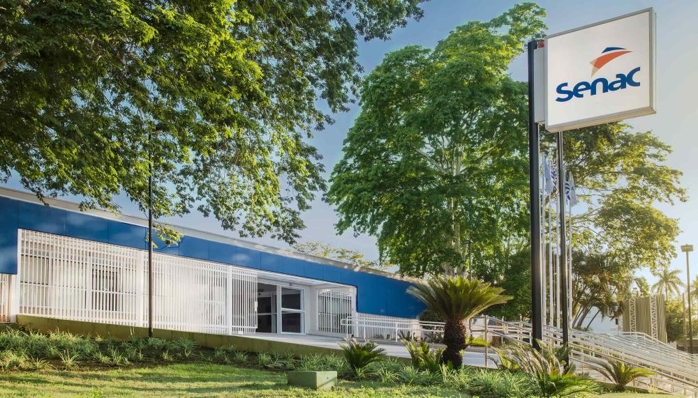 Foto da fachada da unidade. Ela tem a cor azul médio, com grades brancas. Em primeiro plano há um jandim gramado, com plantas rasteiras e médias. Na frente do prédio, grandes árvores projetam sua sombra. Há um letreiro com a logo do Senac no alto.