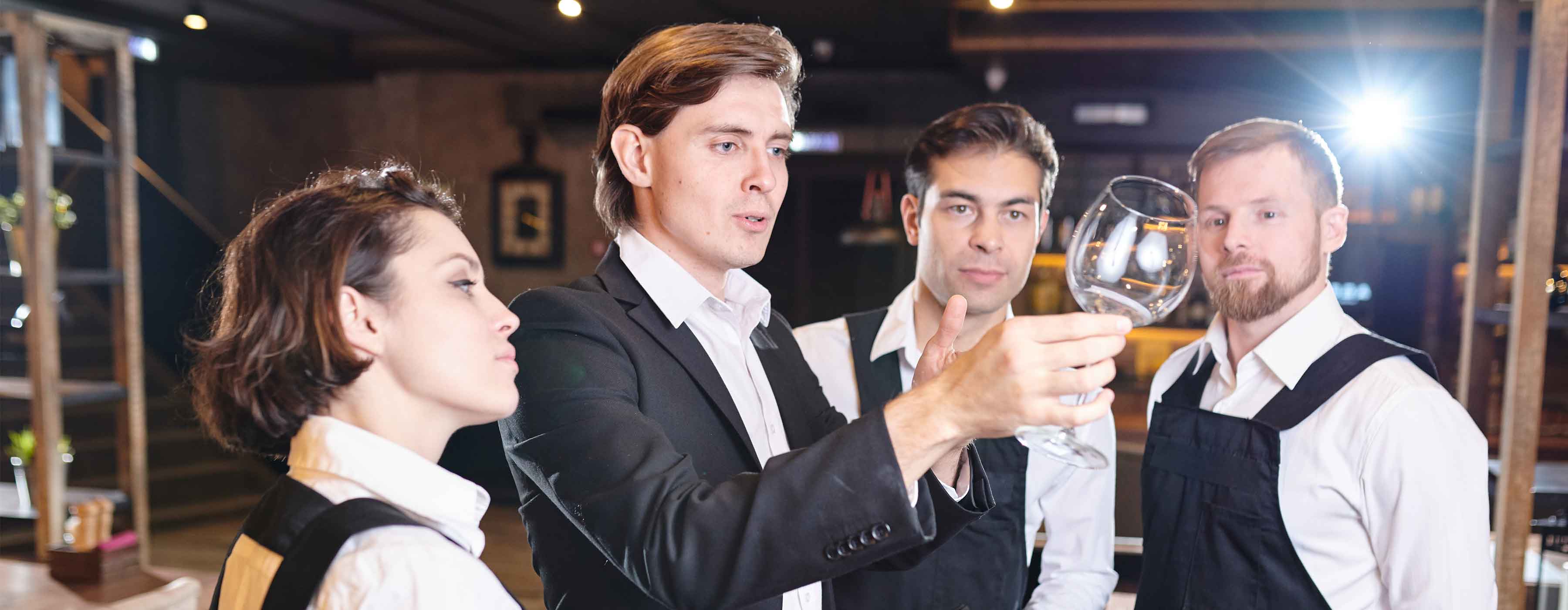 Maître: supervisão de salão - Foto de um maître segurando uma taça, com mais três atendentes de salão