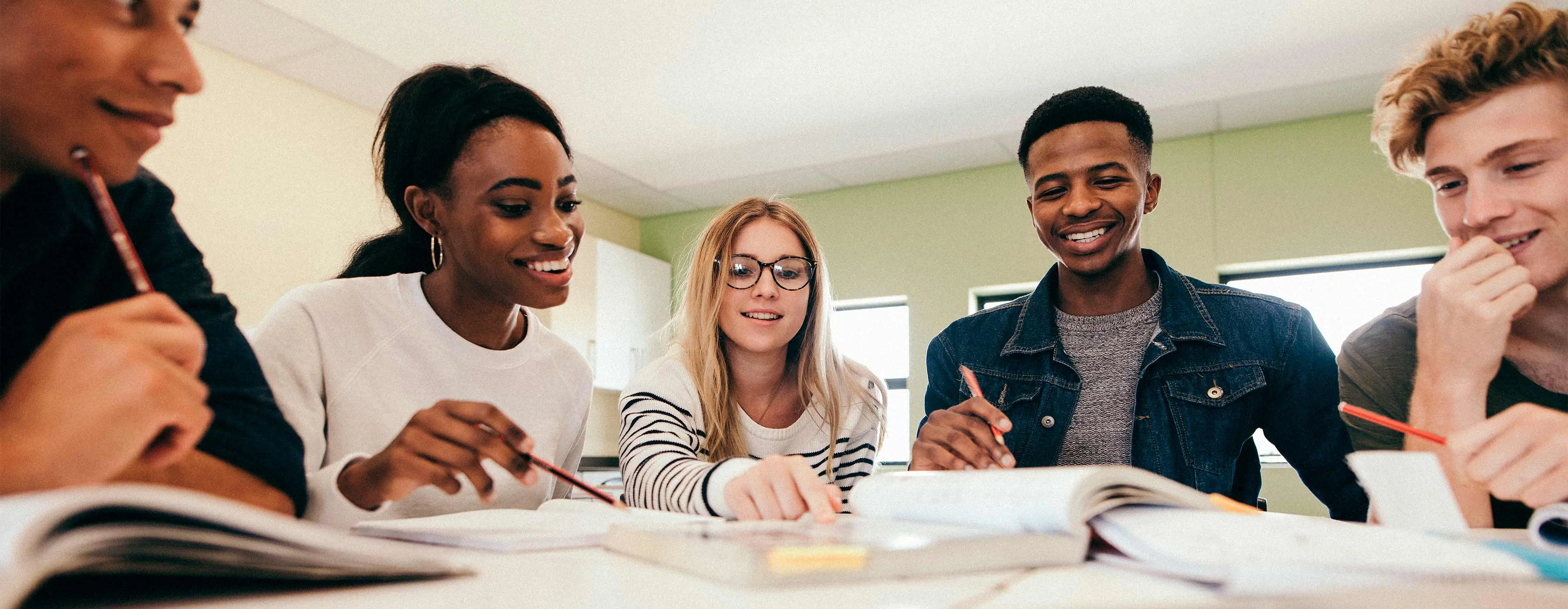 Foto de cinco jovens sorridentes conversando ao redor de um livro sobre uma mesa de estudo.