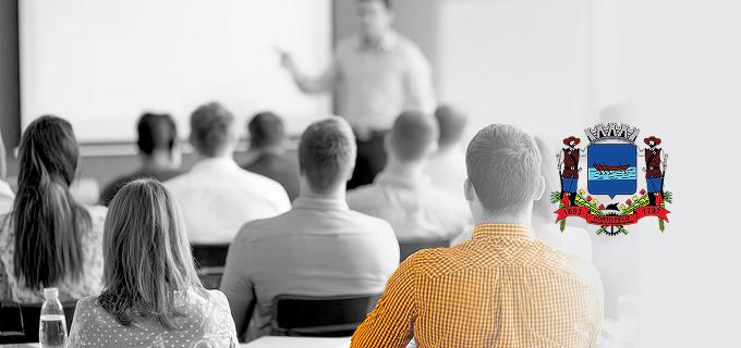 Foto de sala de aula com pessoas sentadas de costas, assistindo a uma apresentação de uma pessoa ao fundo. Ao lado direito, aparece o brasão oficial da cidade de Porto Feliz.