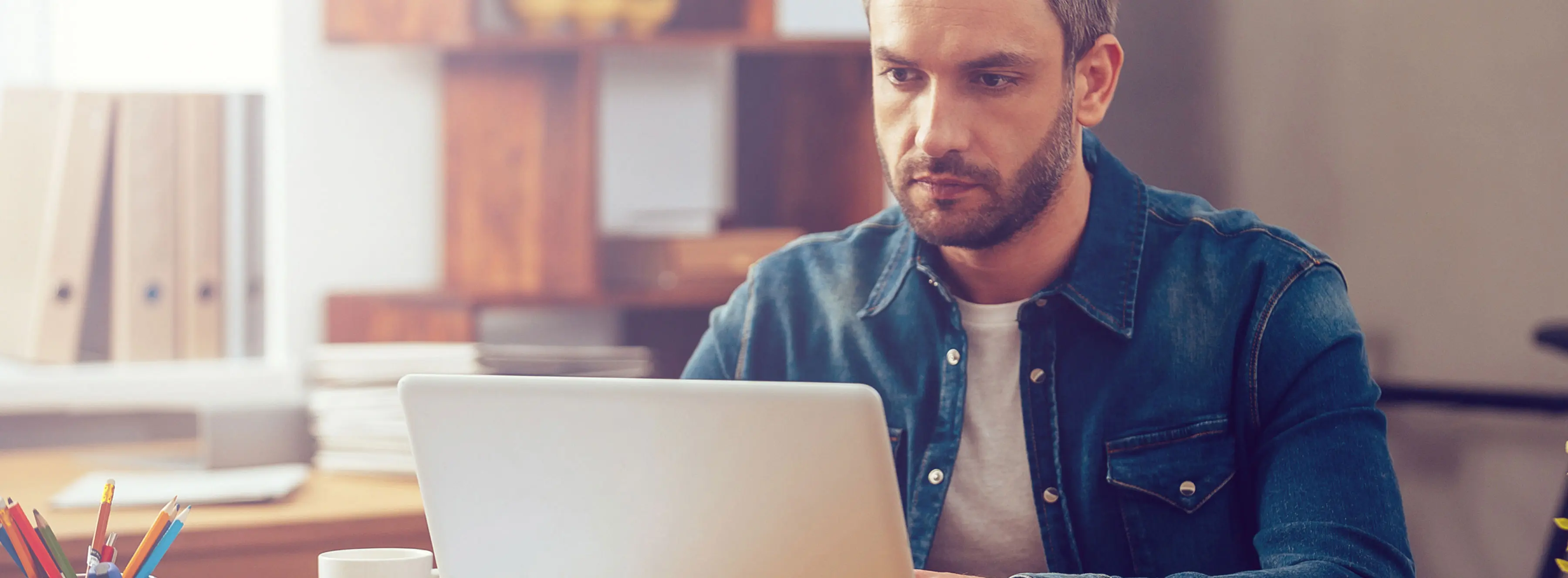 Foto mostra pessoa trabalhando em um notebook sobre uma mesa, com prateleiras ao lado.