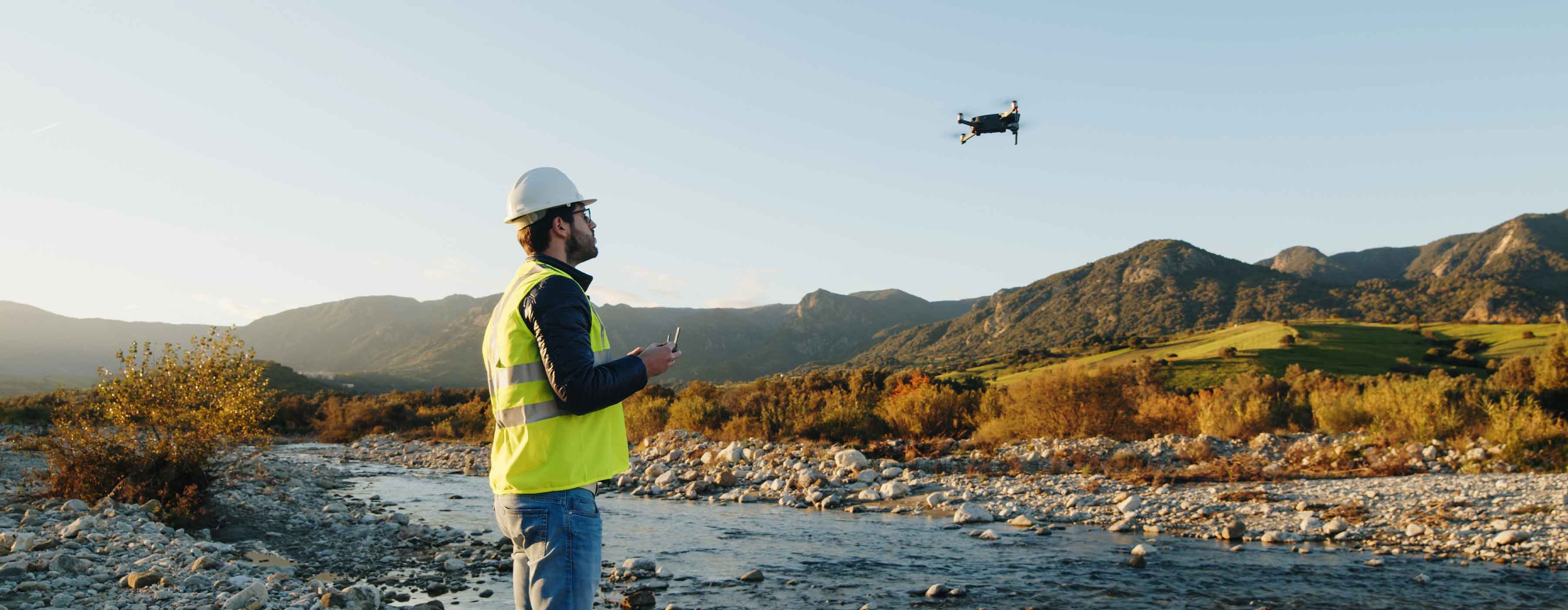 Homem em pé próximo a um rio com pedras, operando um drone com controle remoto, enquanto o equipamento voa no céu, tendo montanhas ao fundo.