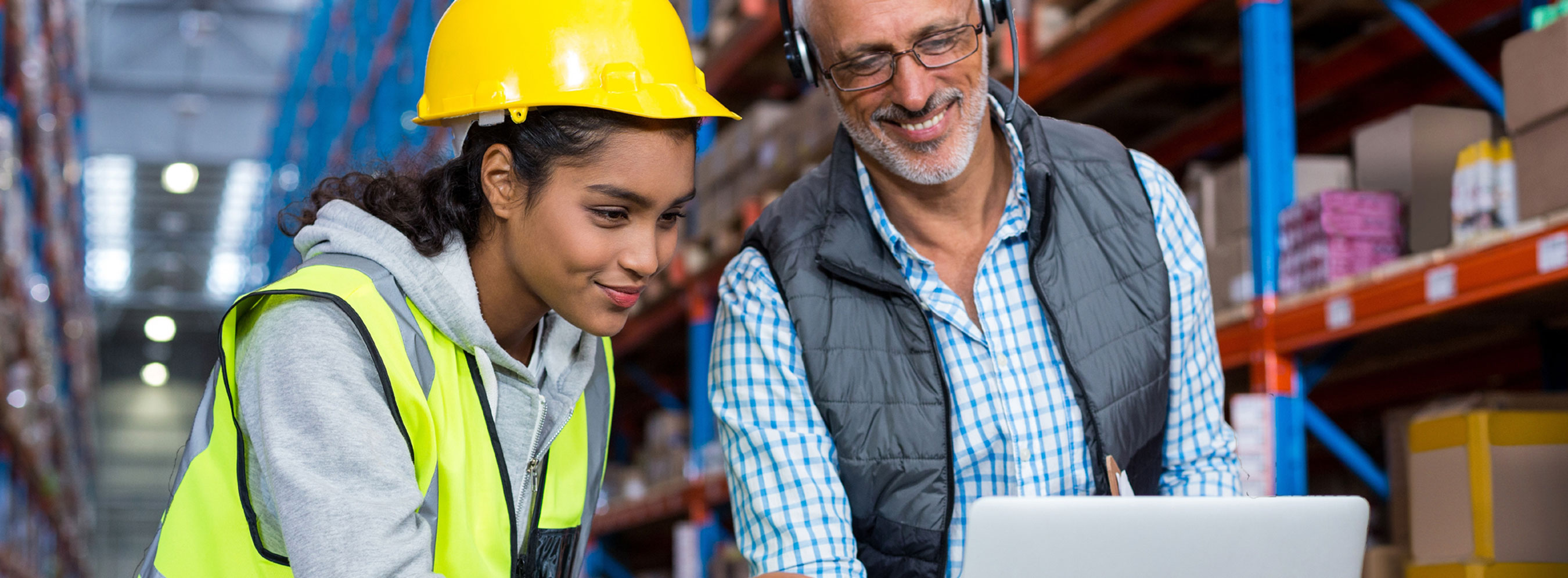 Foto de uma moça com capacete e colete de proteção prestando atenção na tela de notebook, segurada por homem usando fone de ouvido. Eles estão em galpão de logística de empresa.