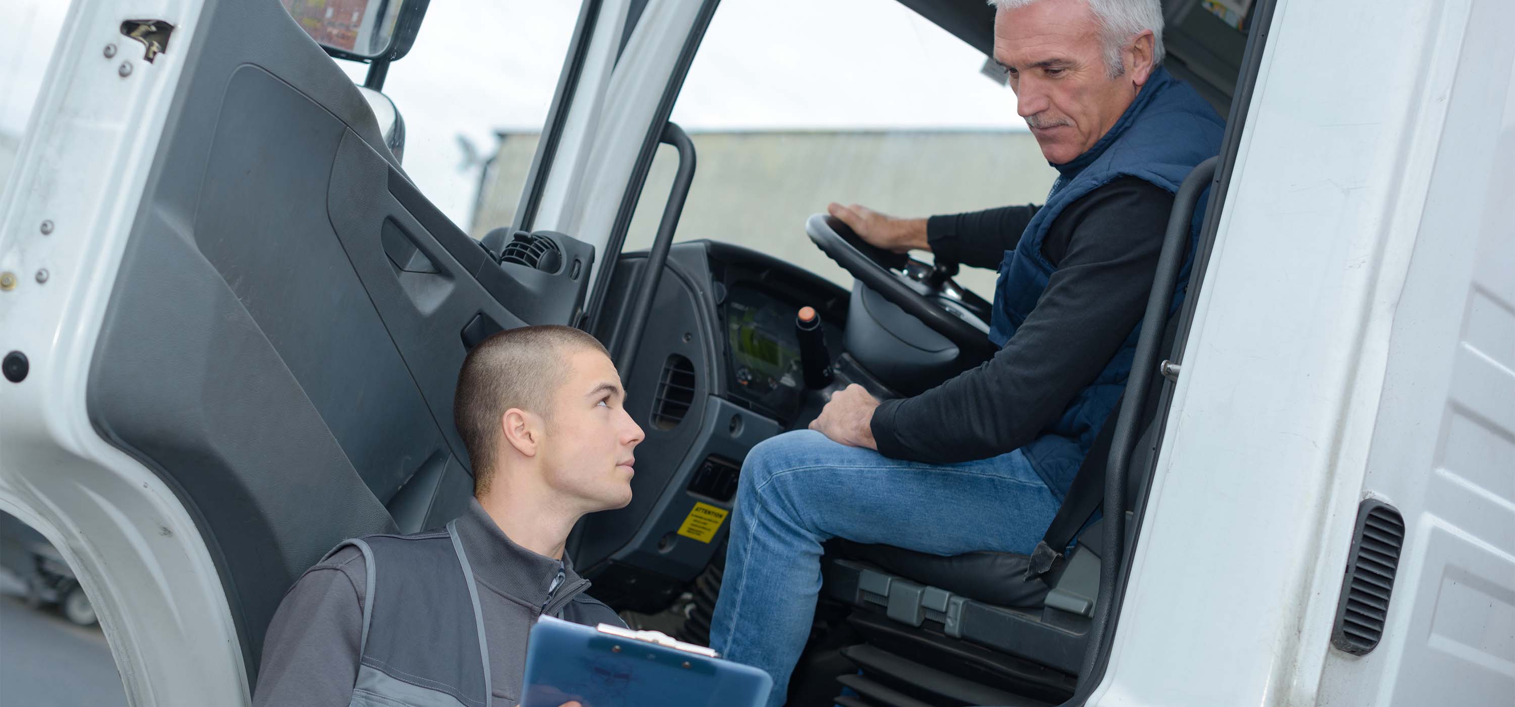 Logística de Transportes. Foto de um homem em um caminhão olhando para uma prancheta mostrada por outro homem, ao lado do veículo.