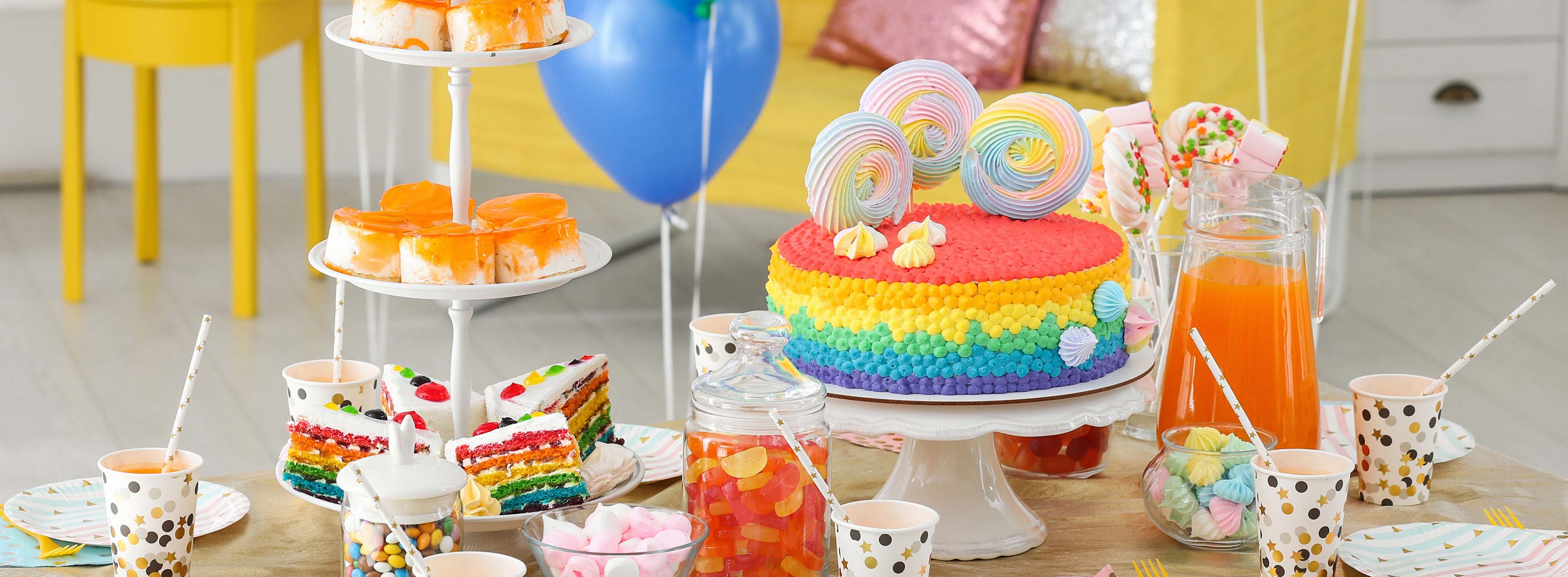Foto de uma mesa em uma festa repleta de doces coloridos, como bolo, suspiros, bala de goma, copos com sucos e algumas bexigas enfeitando o ambiente.
