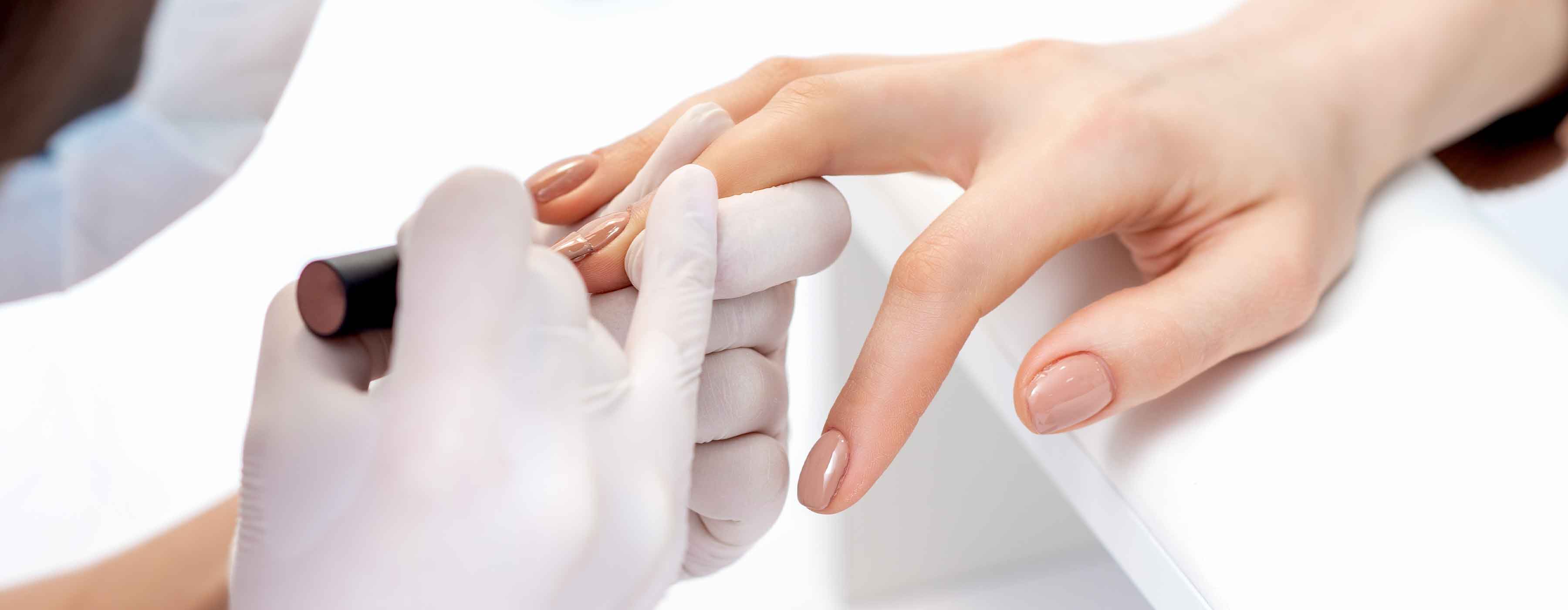 Foto mostra profissional, com luvas brancas passando esmalte cor nude nas unhas de uma cliente.