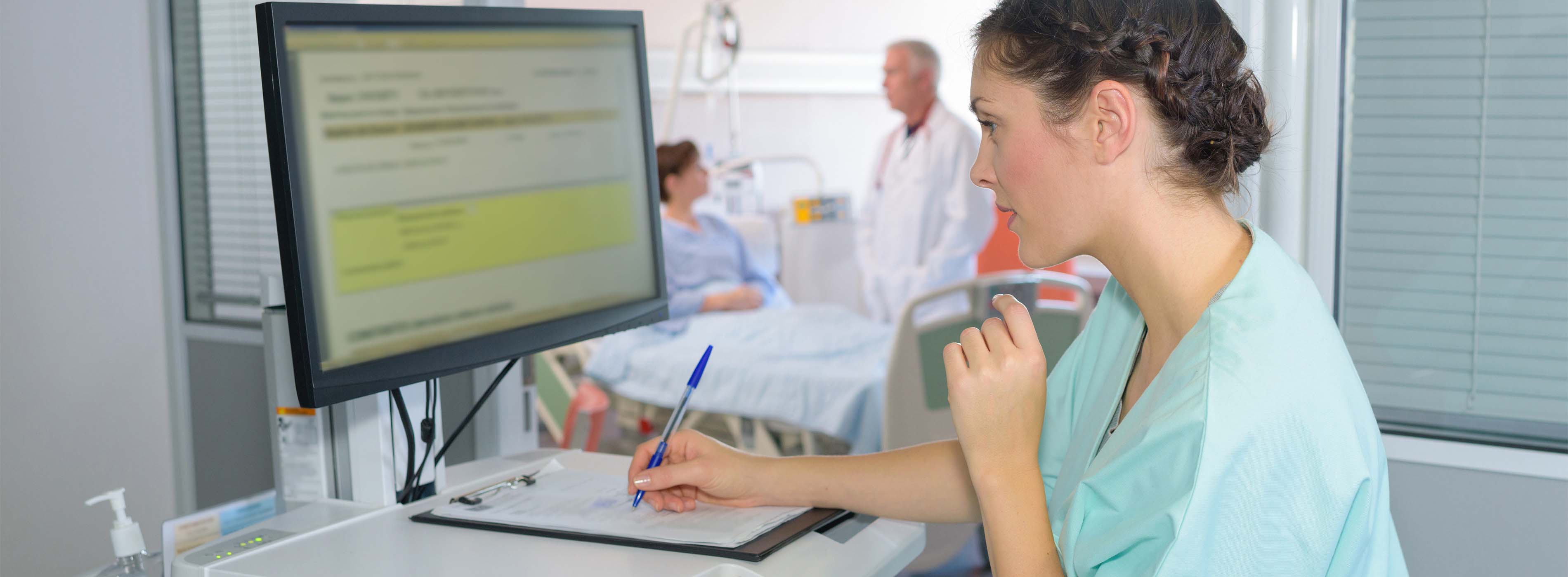 Prontuário Clínico: manual e eletrônico. Foto de mulher com uniforme hospitalar, vendo dados em um sistema de computador e anotando em um papel. Ao fundo, o cenário é de hospital.