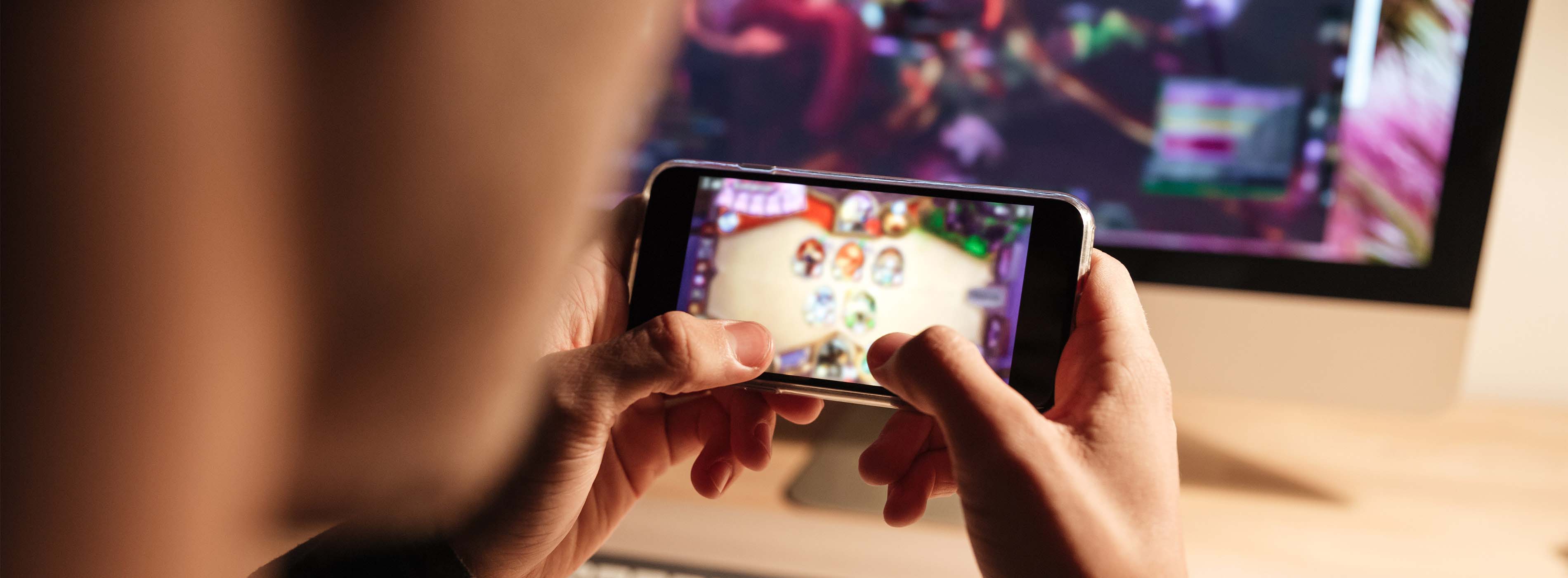 Games 2D - jogos para dispositivos móveis - foto de uma pessoa brincando em uma game em celular e ao fundo tela de computador.