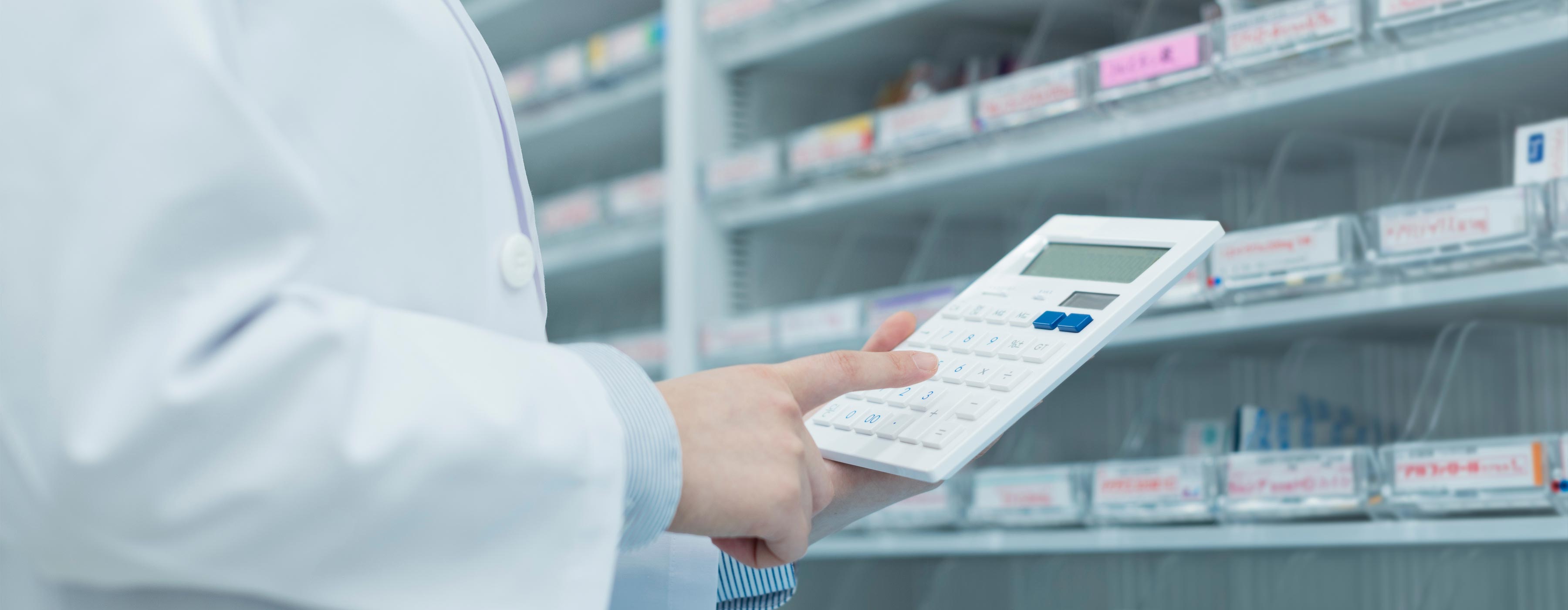 Cálculos Utilizados na Dispensação e Controle de Estoque de Medicamentos - Foto das mãos de uma pessoa utilizando calculadora em frente a uma prateleira com medicamentos.