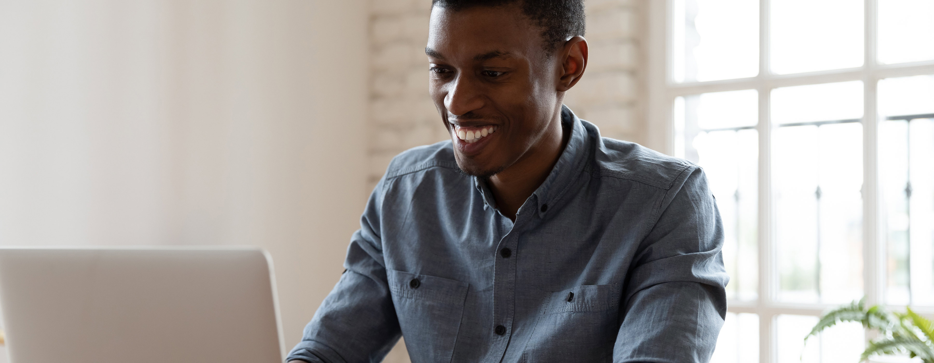 Foto de um homem jovem sorridente, sentado trabalhando em um escritório e digitando em um notebook.
