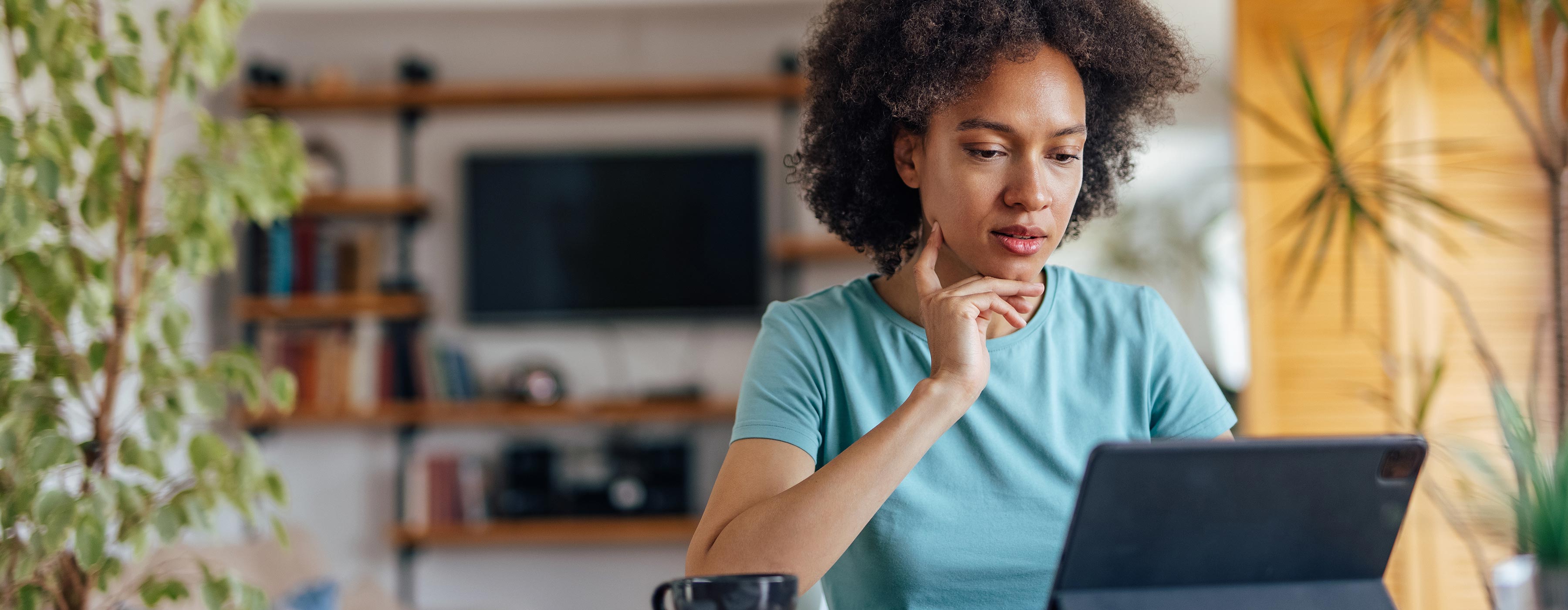 Foto de uma mulher jovem sentada em sua casa, trabalhando home office, verificando atenciosamente informações em um tablet em cima da mesa.
