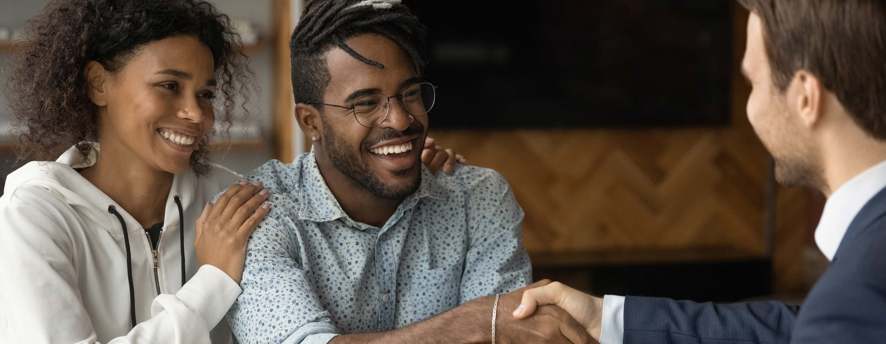 Foto de um homem e uma mulher sorridentes, formando um casal jovem, onde ele aperta as mãos de um outro homem, como sinal de fechamento de acordo em um escritório.
