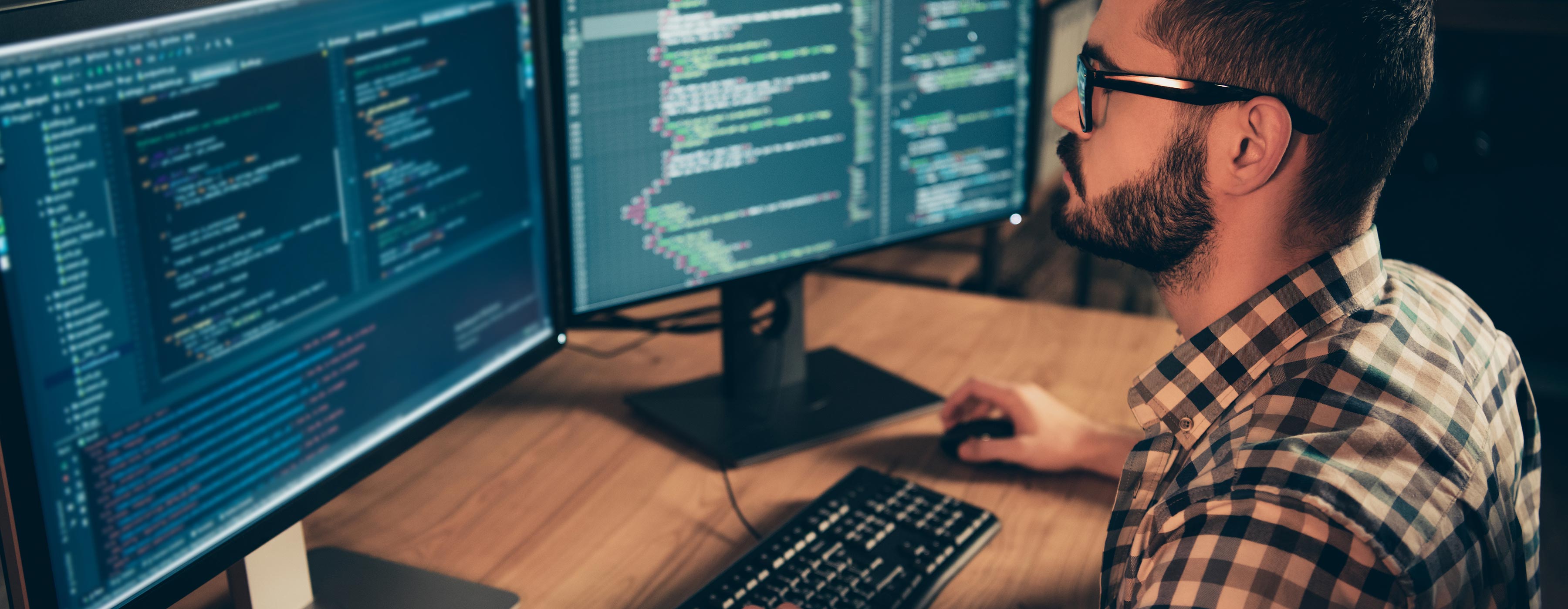 Foto em close da lateral de um homem hovem digitando códigos de sistemas tecnológicos de frente para dois monitores em uma mesa de escritório.