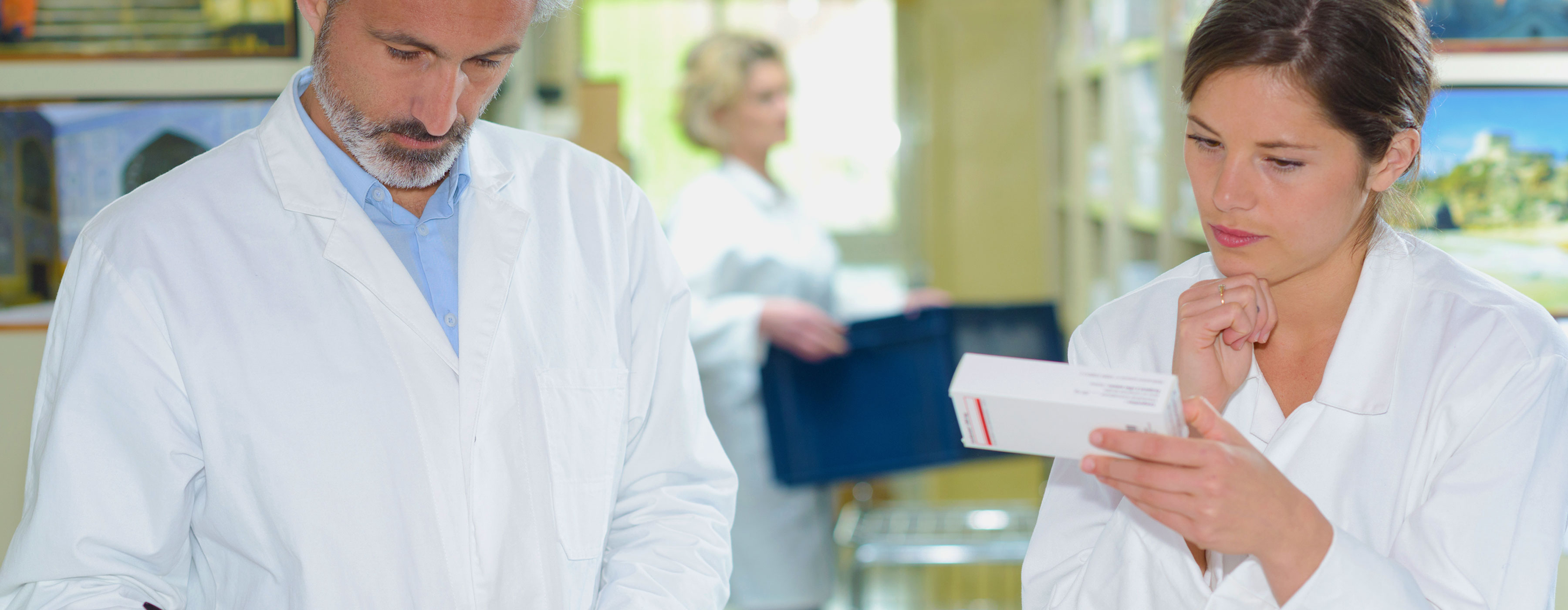 Farmácia Hospitalar: conhecendo os materiais médico-hospitalares. Foto de um homem e uma mulher, vestidos com avental branco, verificando embalagens de remédios em uma farmácia.