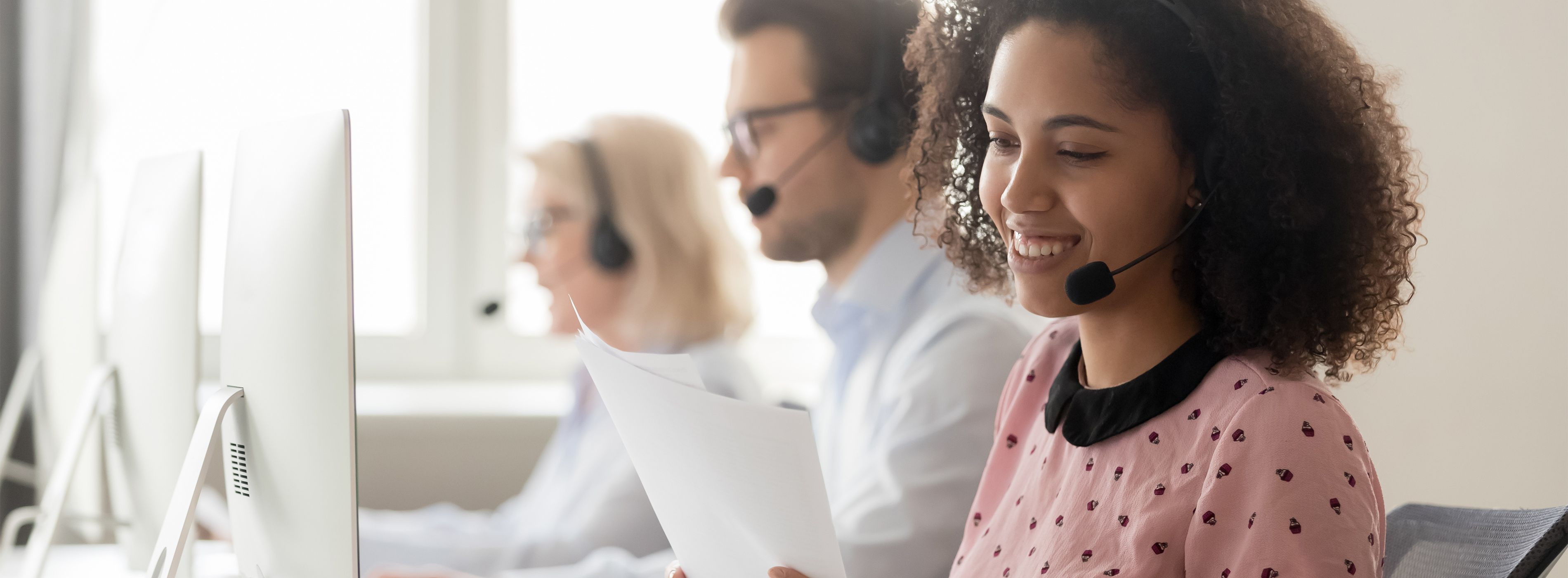 Telemarketing: atendimento ao cliente e rotinas operacionais - foto de mulher usando fone de ouvidos que tem microfone enquanto segura uma folha em frente a um computador