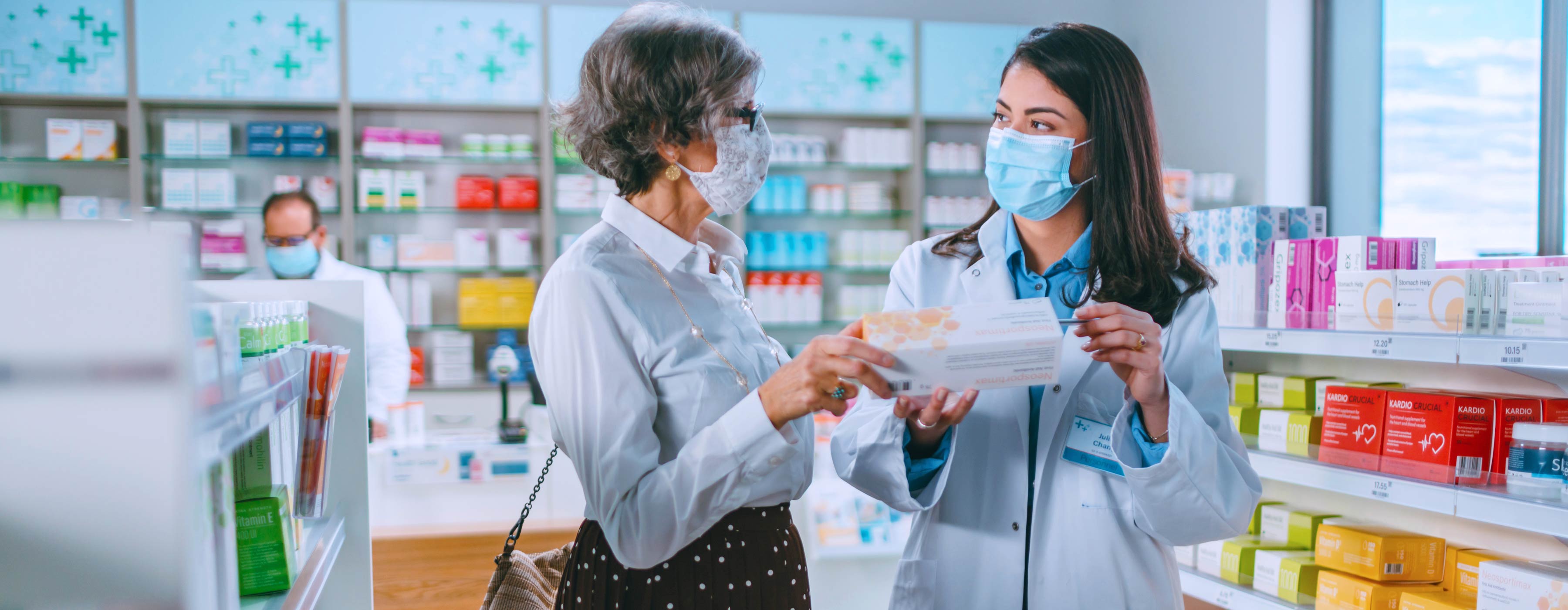 Orientação na Dispensação de Vitaminas, Minerais e Suplementos Alimentares em Drogarias - foto de duas mulheres na farmácia. Uma segura remédio e consulta funcionária do negócio, ambas usam máscaras.