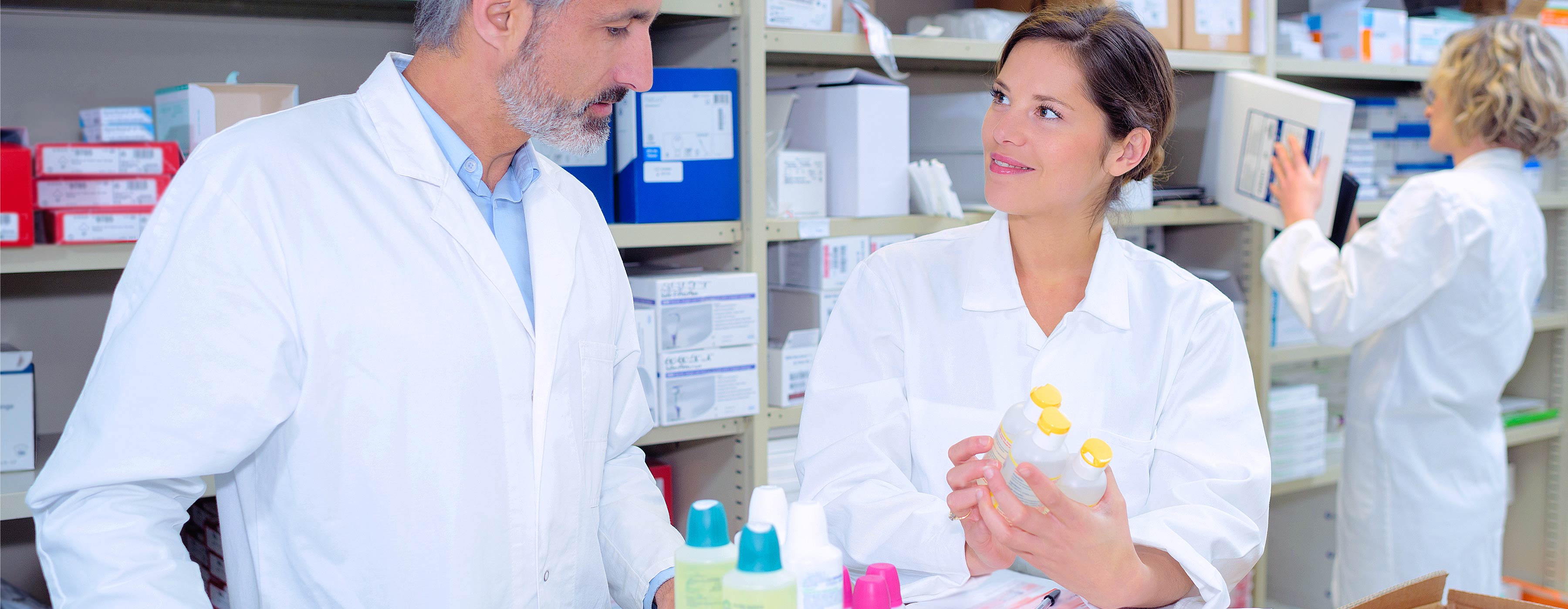 Entendendo a Dispensação na Farmácia Hospitalar. Foto de duas pessoas com avental branco observando recipientes em estoque farmacêutico. Ao fundo, uma moça guarda na estante arquivo.