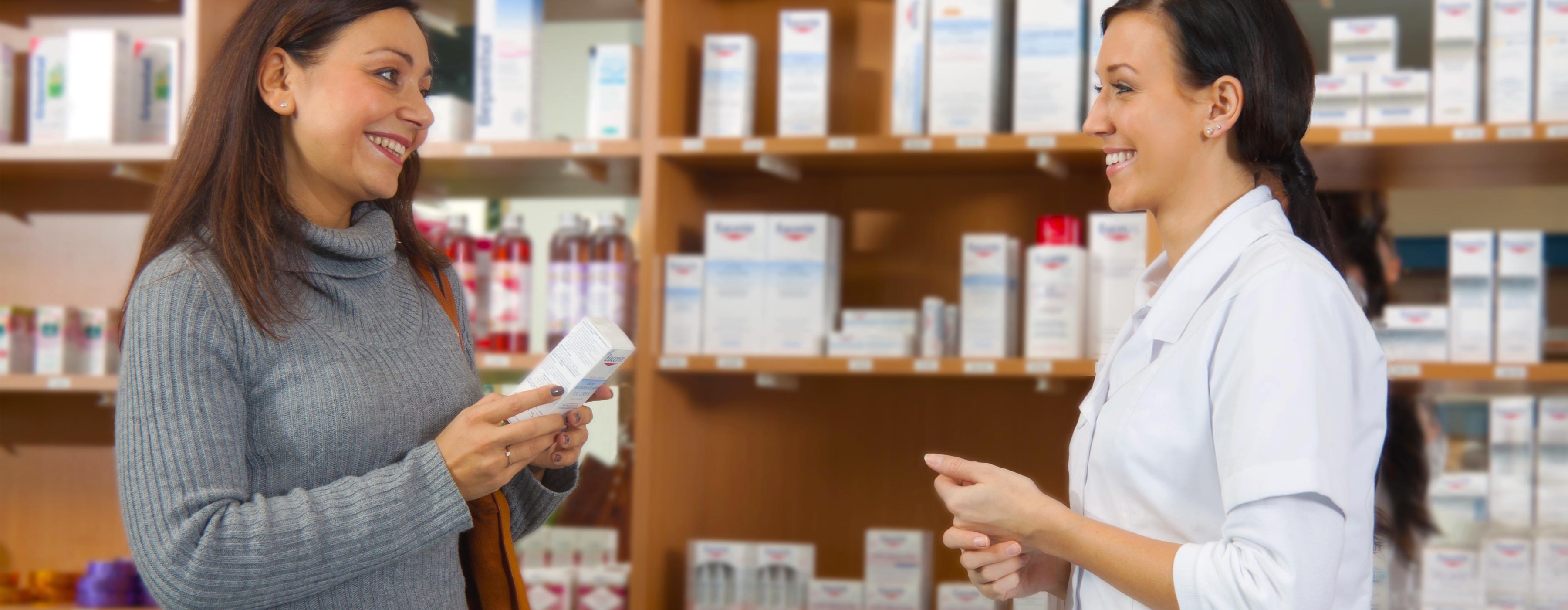 Dermocosmético: orientações para o uso e tendências. Foto de duas mulheres sorrindo em farmácia. Uma usa avental e outra segura uma pequena caixa, ao fundo estante de produtos dermatológicos.