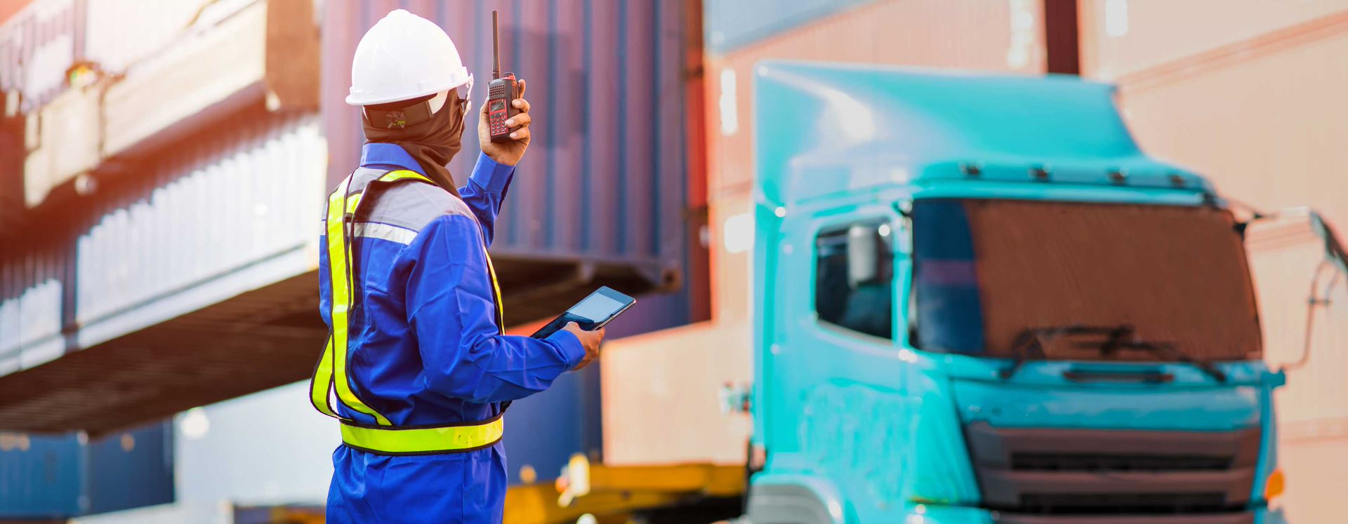 Roteirização de cargas - foto de Profissional de Logística vestindo EPI observa caminhão de container manobrando enquanto dá diretrizes através do rádio