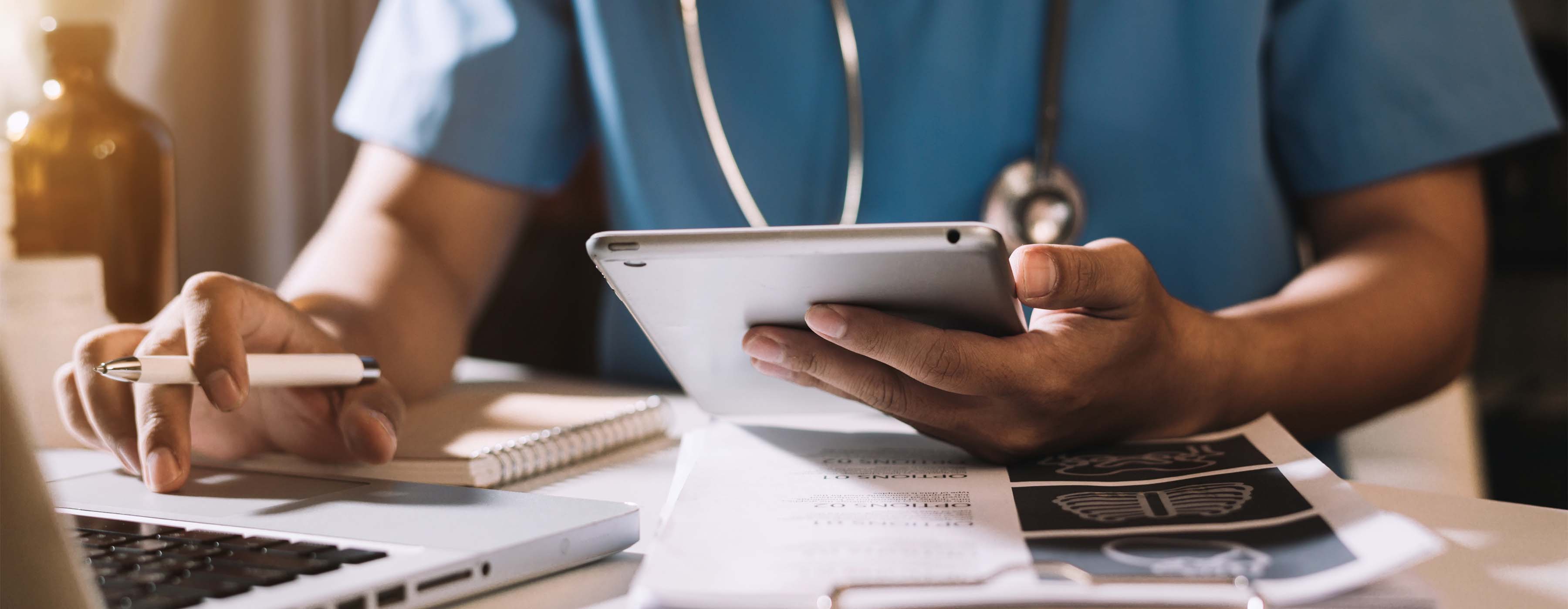 Gestão de Compras e Estratégias de Negociação em Serviços de Saúde. Foto de profissional de saúde sentado em frente à mesa de consultório com estetoscópio no pescoço e, em uma das mãos, um tablet, e na outra, caneta entre os dedos enquanto digita no notebook