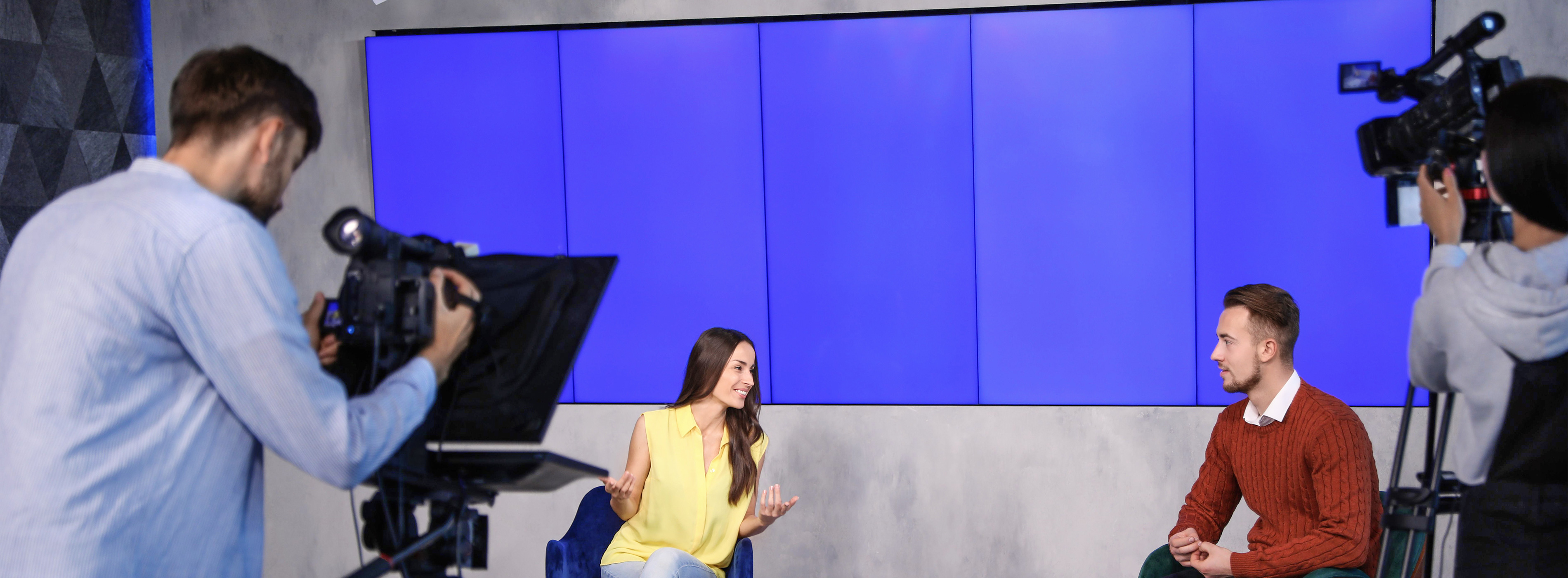 Radialista Apresentação de Programas de Televisão. Foto de dois operadores de câmera filmando um homem e uma mulher sentados que conversam em cenário de programa de televisão.