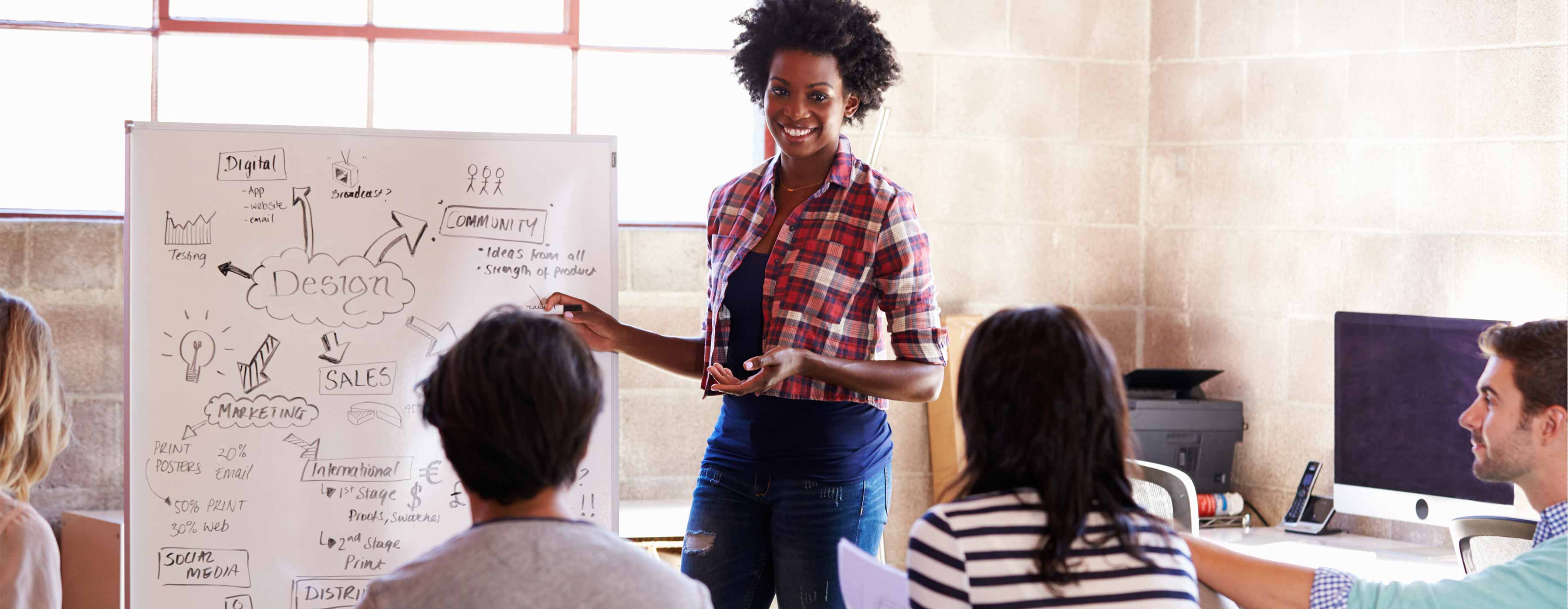 Facilitação Gráfica: Resumo visual para comunicar ideias em reuniões e palestras - Foto de uma mulher negra, sorrindo e apontando para um quadro de anotações enquanto fala com um grupo de pessoas sentadas.