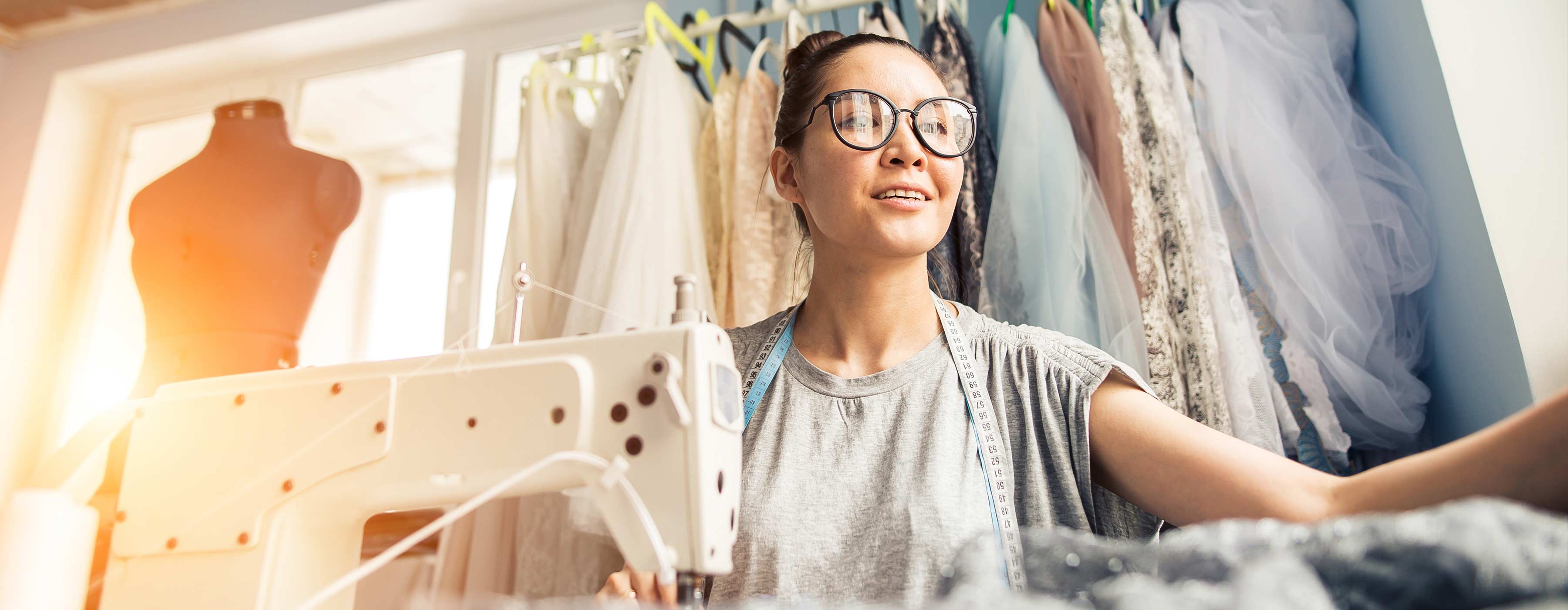 Técnicas de Acabamentos em Tecidos Finos - Foto em close de mulher jovem em um ateliê de moda utilizando a máquina de costura. Ao fundo, uma arara com roupas e um manequim.