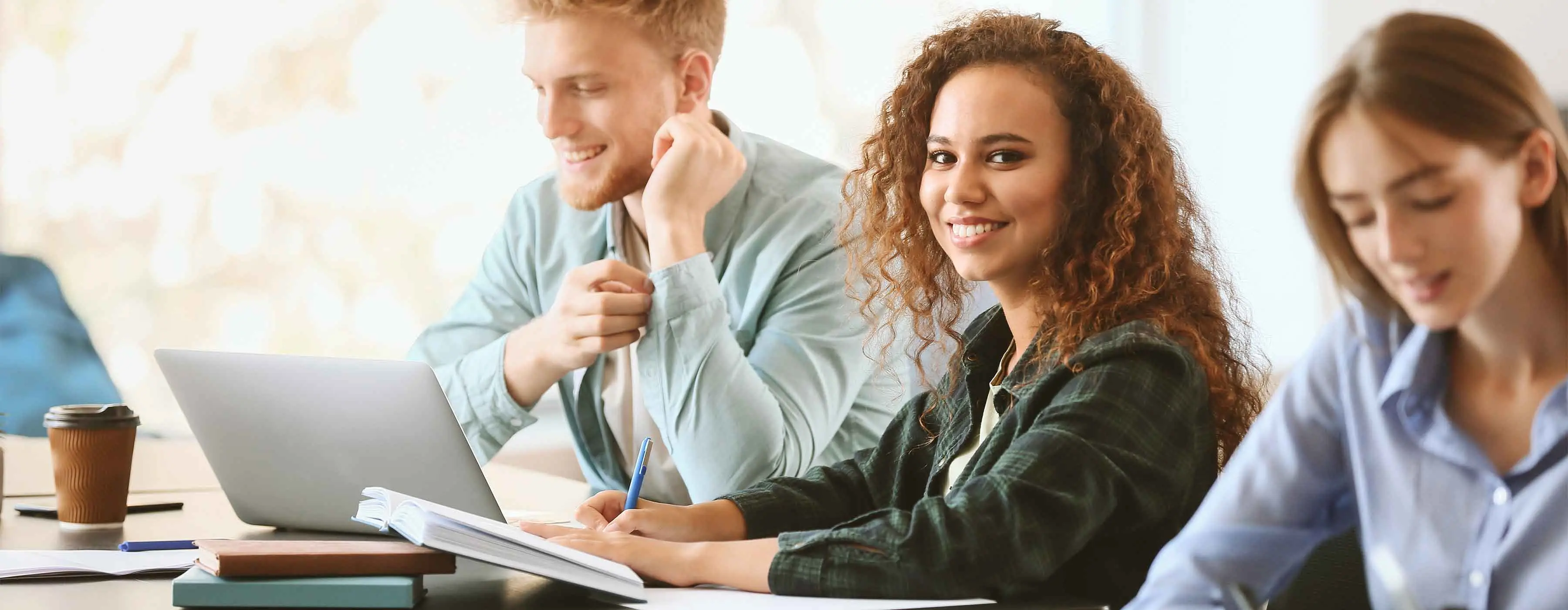 Inglês Avançado. Foto de uma jovem sorridente fazendo anotação em caderno e posando para foto com outras duas pessoas ao lado dela.