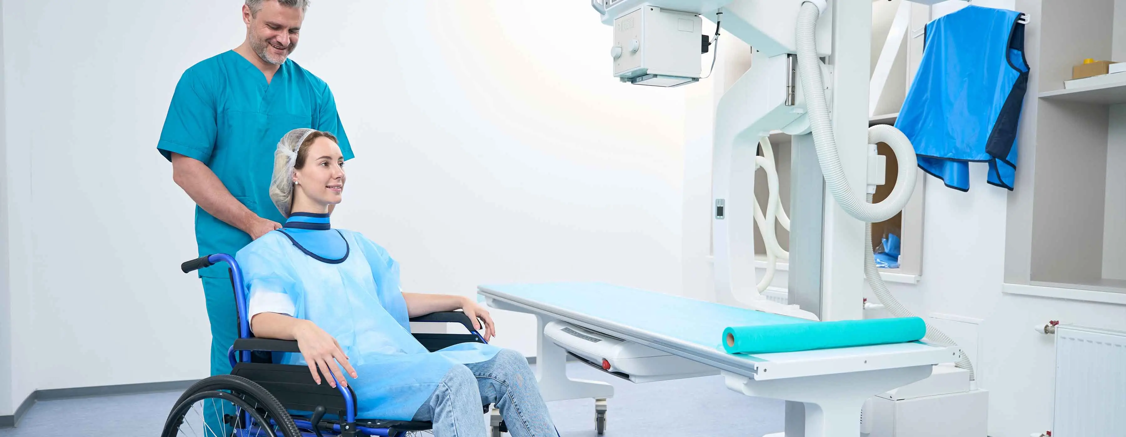 Proteção e Segurança no Radiodiagnóstico - foto de mulher na cadeira de rodas sendo deslocada por um homem, os dois estão próximos a uma máquina de exames