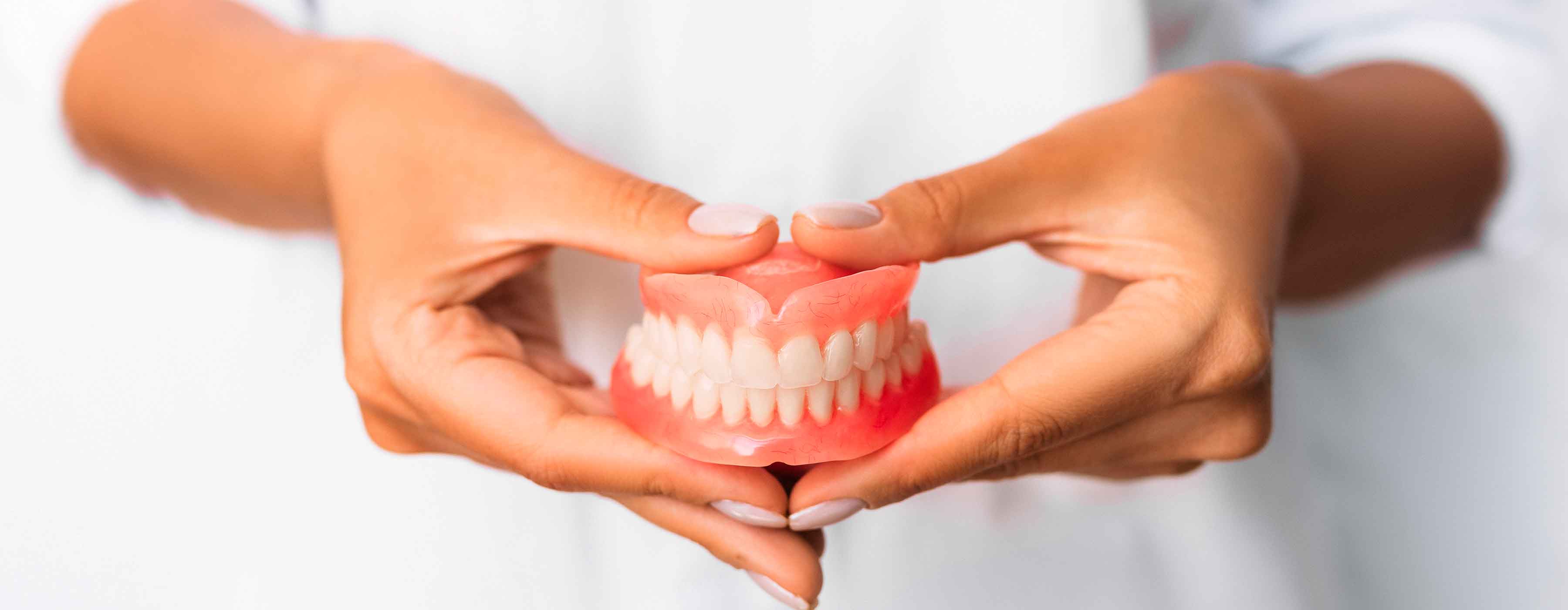 Foto mostra as mãos de uma pessoa segurando uma prótese dentária, com gengiva rosada e dentes brancos.