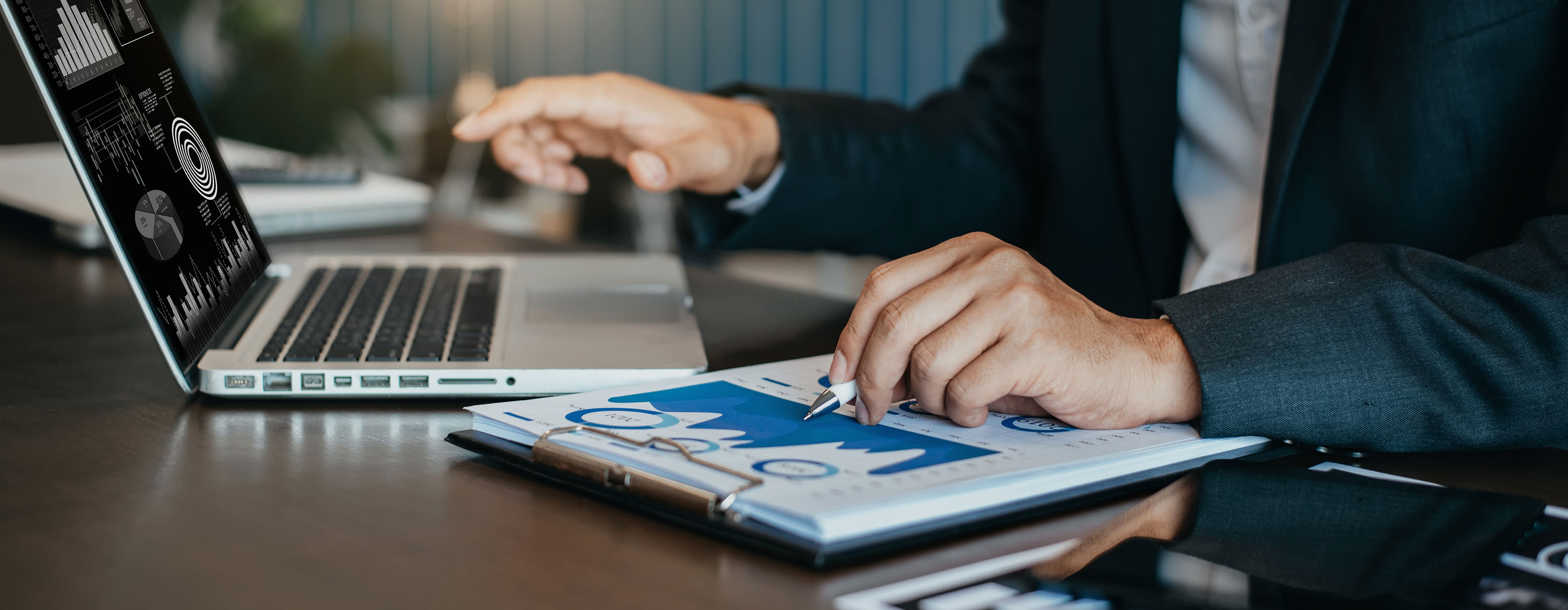 Gestão de Crise nas Redes Sociais - foto em close de uma pessoa mexendo em um notebook e ao mesmo tempo verificando documentos gráficos e financeiros em um caderno em cima da mesa.