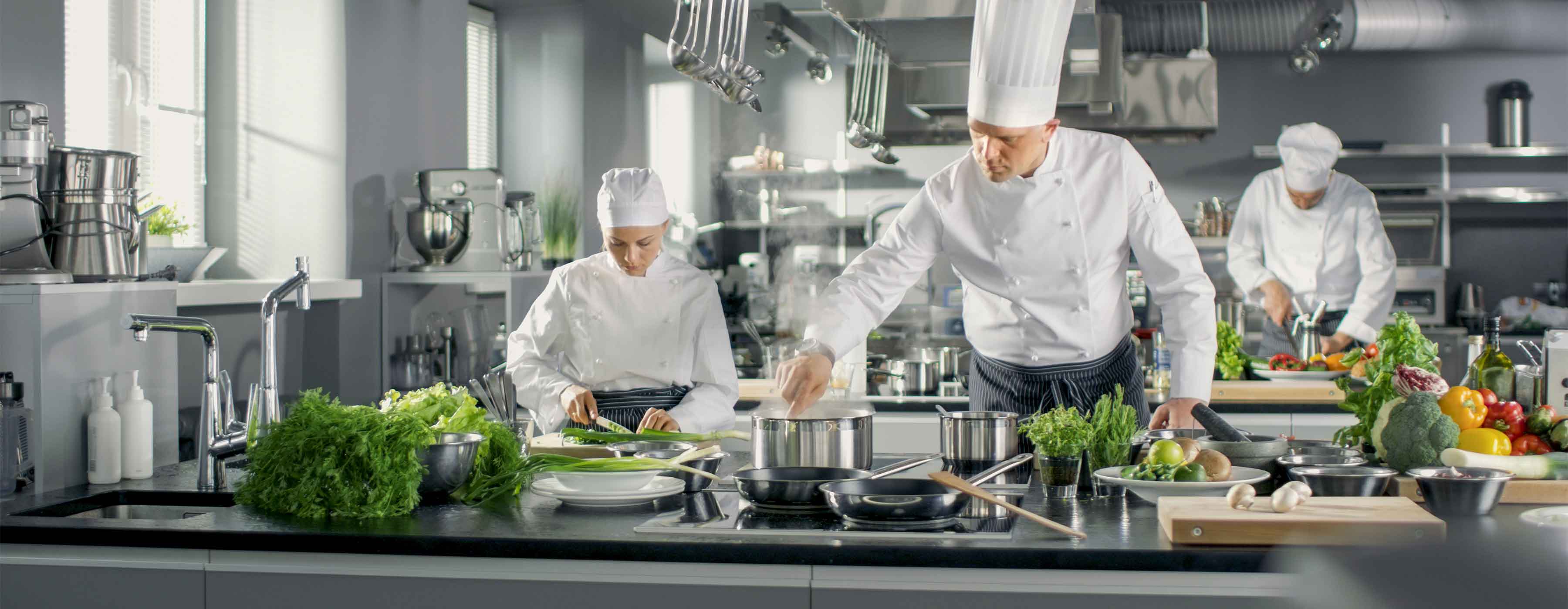 Foto mostra três pessoas cozinhando em bancadas equipadas, com panelas, utensílios, legumes e verduras.