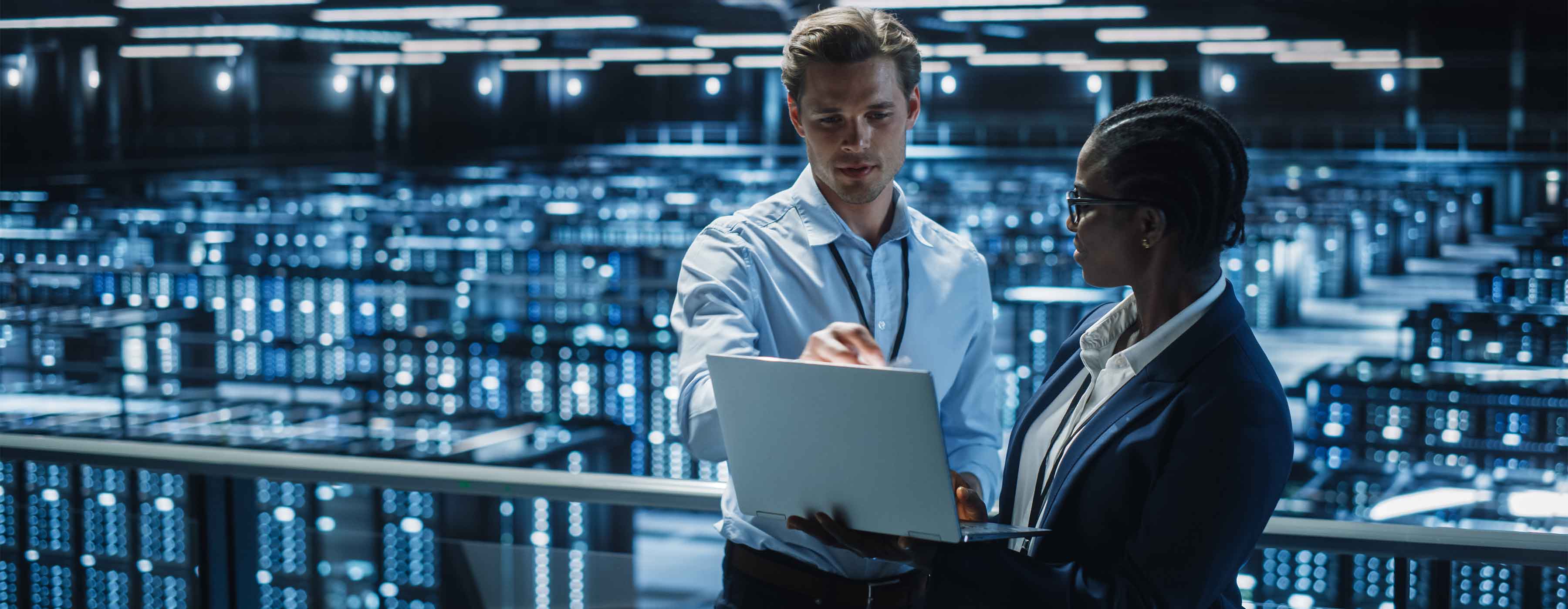 Foto de duas pessoas analisando informações em um notebook, em um grande data center, com racks de servidores iluminados ao fundo.