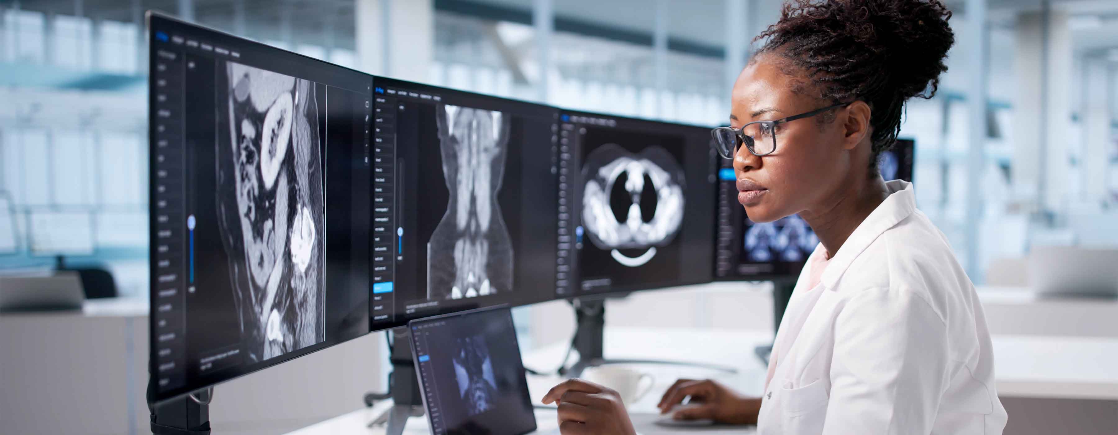 Foto de uma mulher profissional da saúde analisando imagens de exames médicos em três monitores, em ambiente moderno de laboratório.