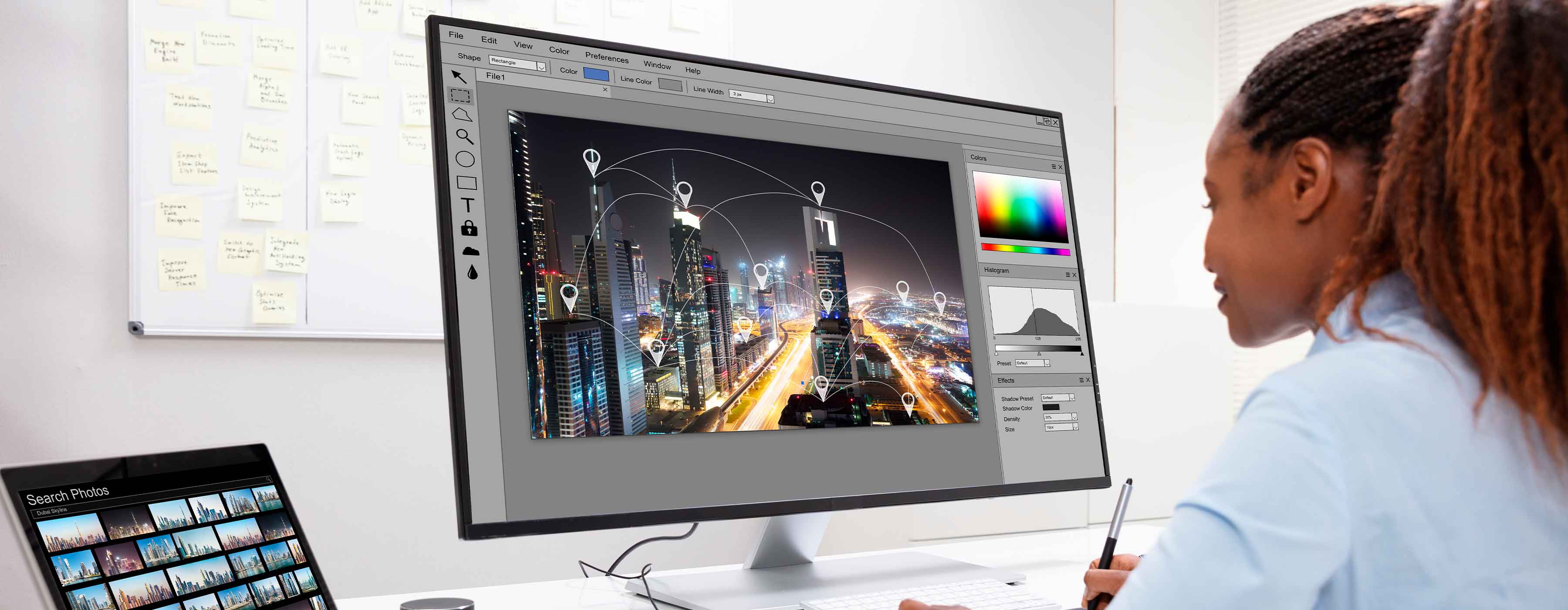 Foto mostra pessoa em frente a um computador editando imagens de uma cidade iluminada com e marcações gráficas.