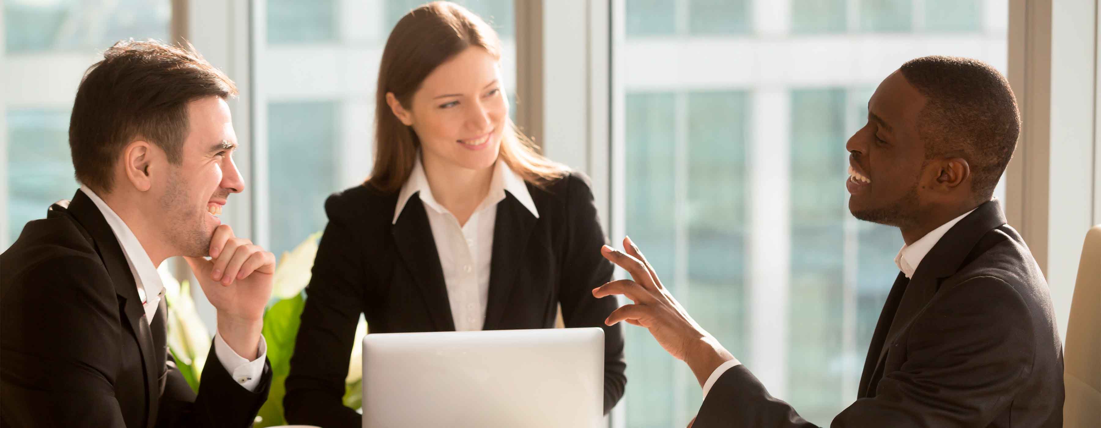 Foto de três pessoas em reunião de negócios, usando trajes sociais, sentadas ao redor de uma mesa com notebook, conversando em ambiente corporativo iluminado por luz natural.
